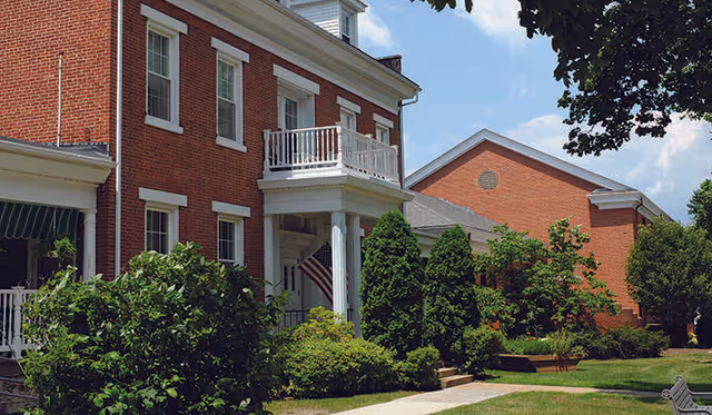 Exterior view of a red brick building with white trim and a small balcony above the entrance. The entrance is flanked by white columns and an American flag is displayed near the door. The building is surrounded by green bushes, trees, and a well-maintained lawn under a partly cloudy sky.