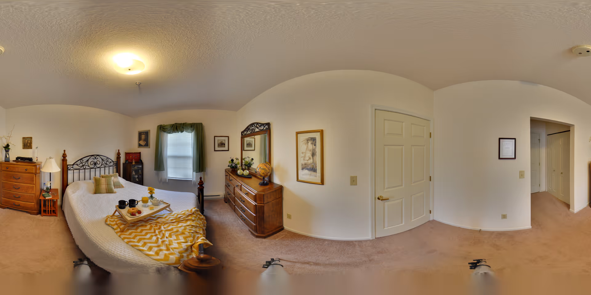 A cozy bedroom with a large bed covered in a white quilt and a yellow patterned throw blanket. There is a wooden dresser with a mirror, a chest of drawers, and a nightstand with a lamp. The room has beige carpet, white walls, a window with green curtains, and framed artwork on the walls.