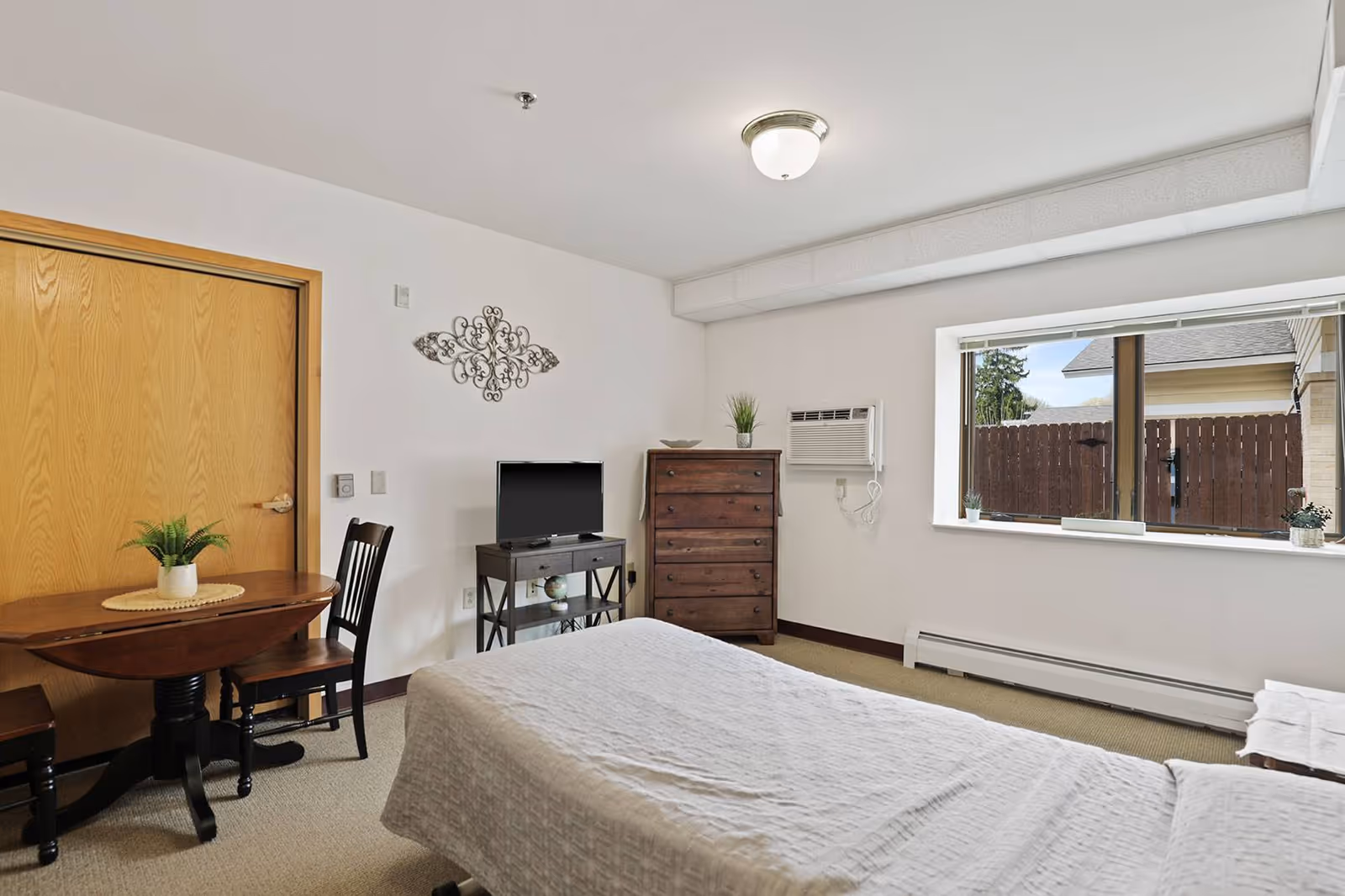 A senior living facility bedroom with a bed covered in a light-colored quilt, a wooden dresser, a small TV on a stand, a round wooden table with two chairs, and a large window showing an outdoor fence and part of a neighboring building. The room has white walls and beige carpet.