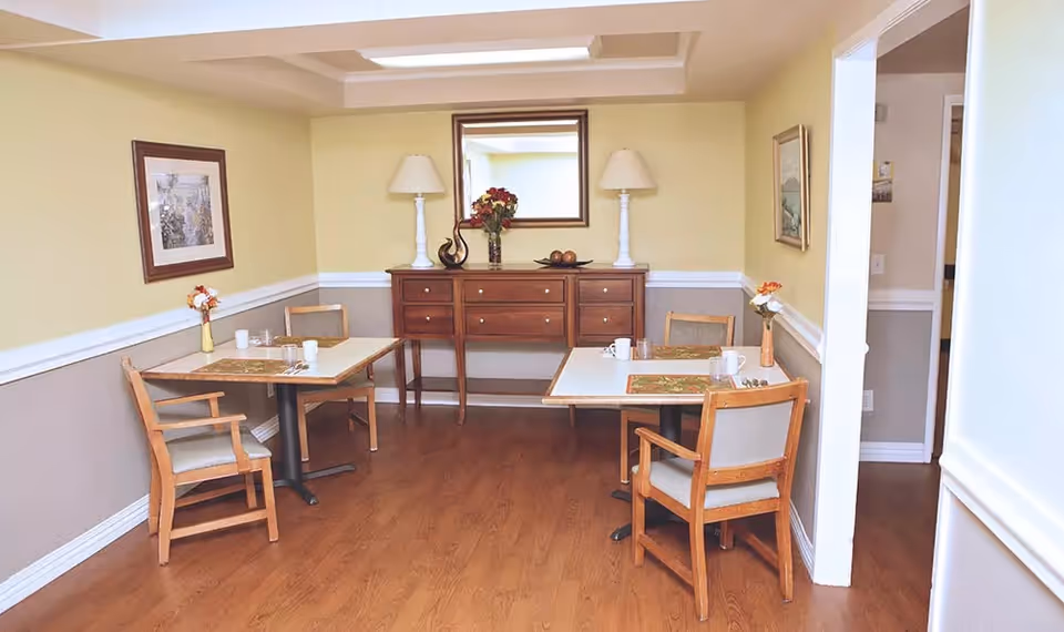 A small dining area with two square tables, each set with placemats, cups, and utensils. Four wooden chairs with cushions are arranged around the tables. The room has light yellow walls with a white chair rail and a wooden floor. A wooden sideboard with two white lamps, a vase with flowers, and decorative items is against the far wall beneath a mirror. Framed artwork hangs on the walls.