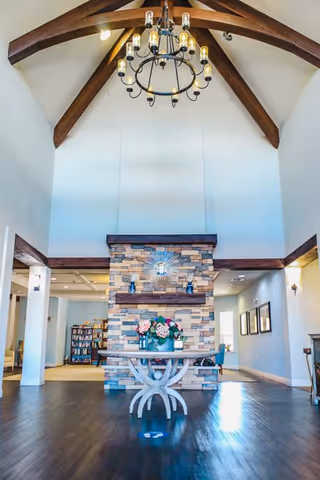 Interior view of a senior living facility common area with a high vaulted ceiling featuring exposed wooden beams and a chandelier. A stone fireplace with a decorative clock and two small statues is centered in the room. In front of the fireplace is a round table with a floral arrangement. Bookshelves and framed pictures are visible in the background.