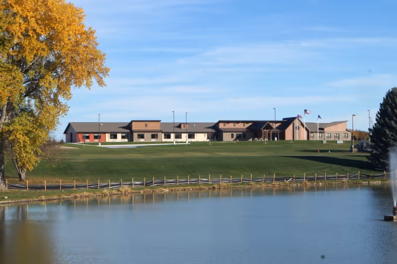 Single-story brick and stucco building with flagpoles on a grassy hill overlooking a pond and fountain, with autumn trees at the edges.