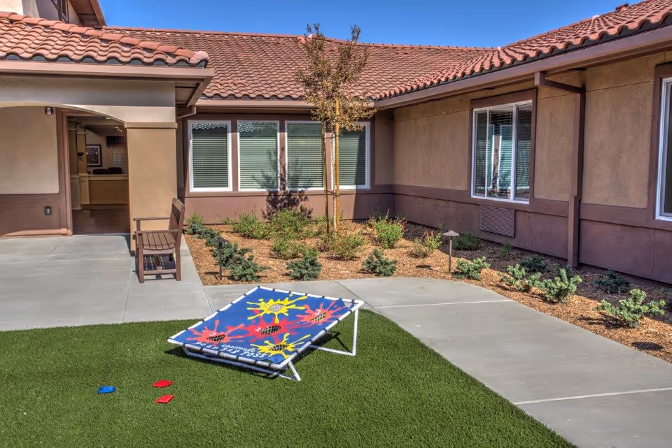 Small outdoor courtyard with artificial turf, a cornhole board and beanbags in front of a single-story building with tiled roof and windows.