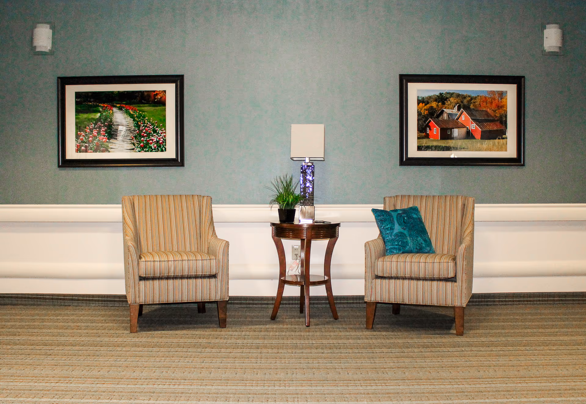 Two striped armchairs flanking a small round table with a lamp and plant against a teal wall with two framed landscape paintings.