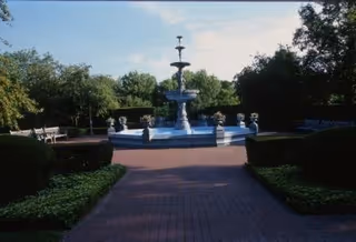 A large outdoor fountain with multiple tiers is centered on a brick pathway, surrounded by neatly trimmed bushes and trees. There are benches placed along the sides of the pathway, and the sky is clear with some clouds.