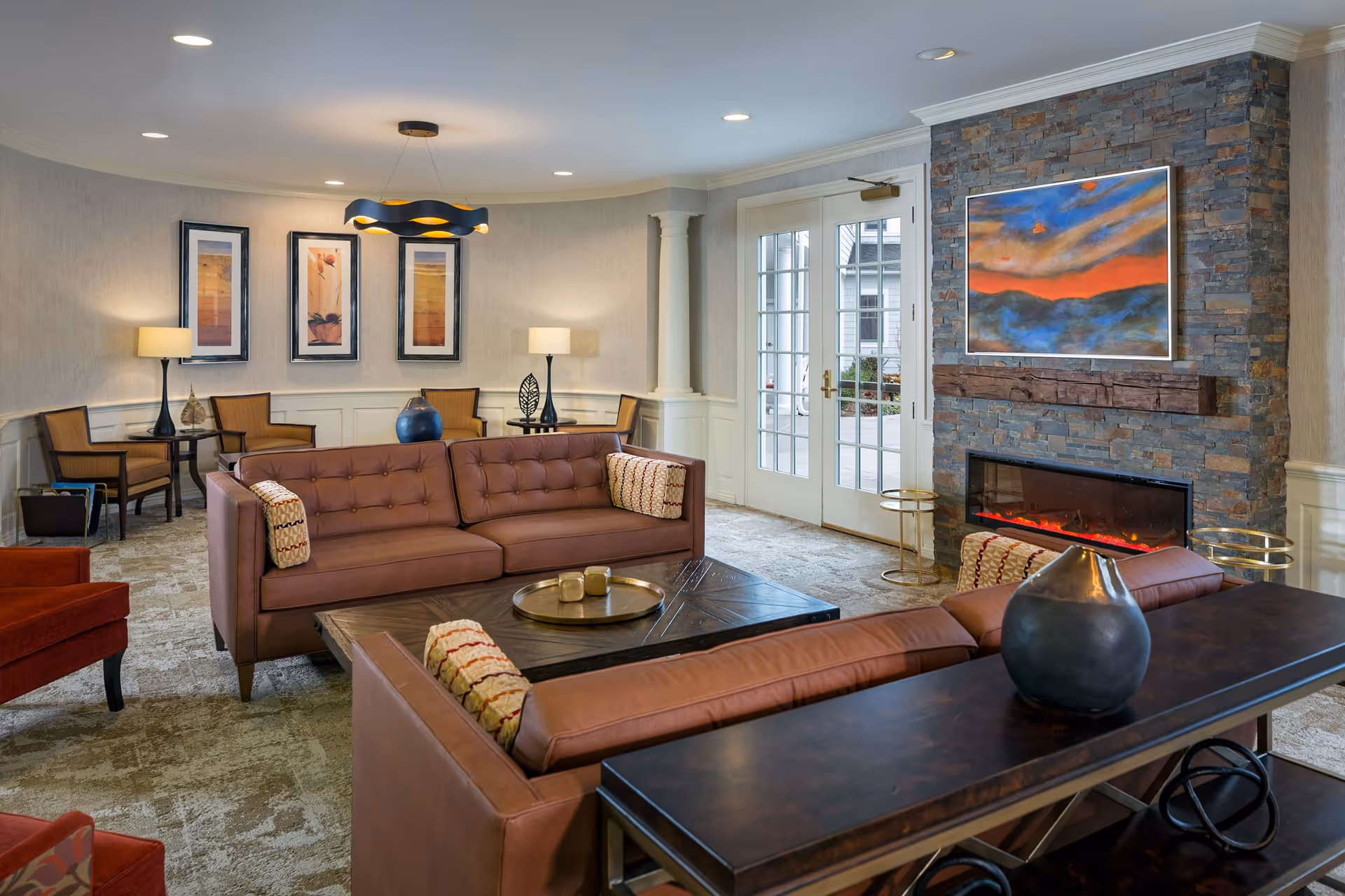 A cozy living room with two brown leather sofas facing each other around a dark wooden coffee table. The room features a stone fireplace with a colorful abstract painting above it, two floor lamps, three framed pictures on the wall, and a modern black ceiling light fixture. French doors lead outside, and the room is carpeted with a patterned design.