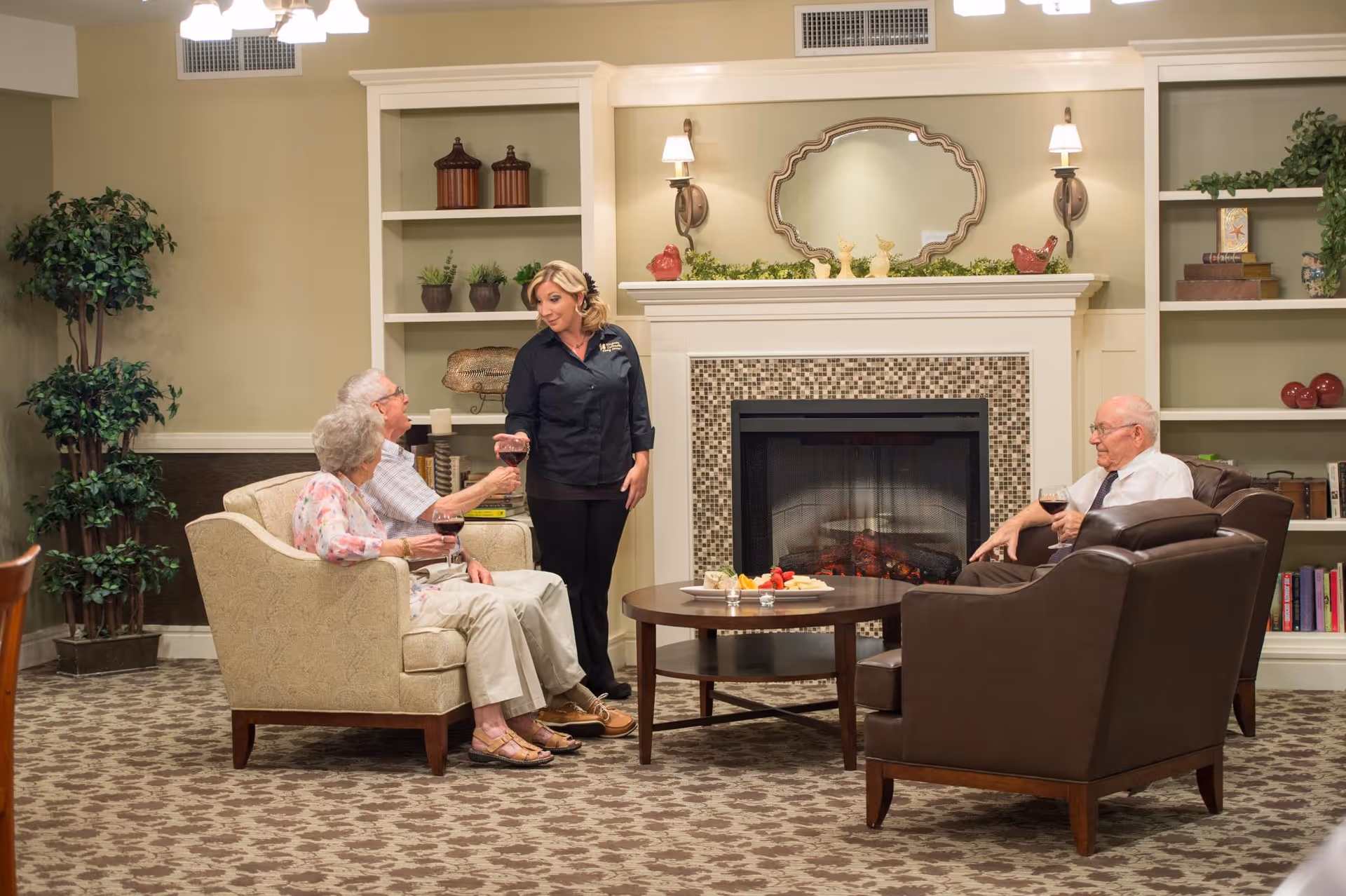 Three elderly residents and a caregiver gathered around a coffee table in a cozy lounge area in front of a fireplace.