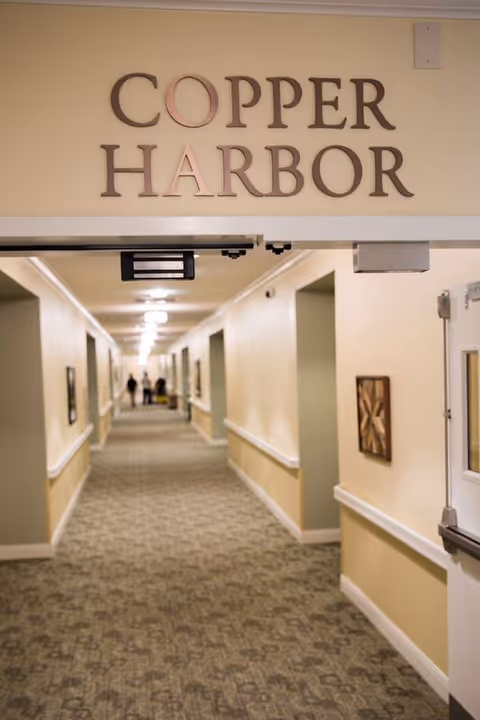 A long, carpeted hallway in a senior living facility with beige walls and handrails on both sides. The hallway is well-lit with ceiling lights and has framed artwork on the walls. Above the hallway entrance, there is a sign that reads 'COPPER HARBOR'. Two people are visible walking in the distance at the end of the hallway.