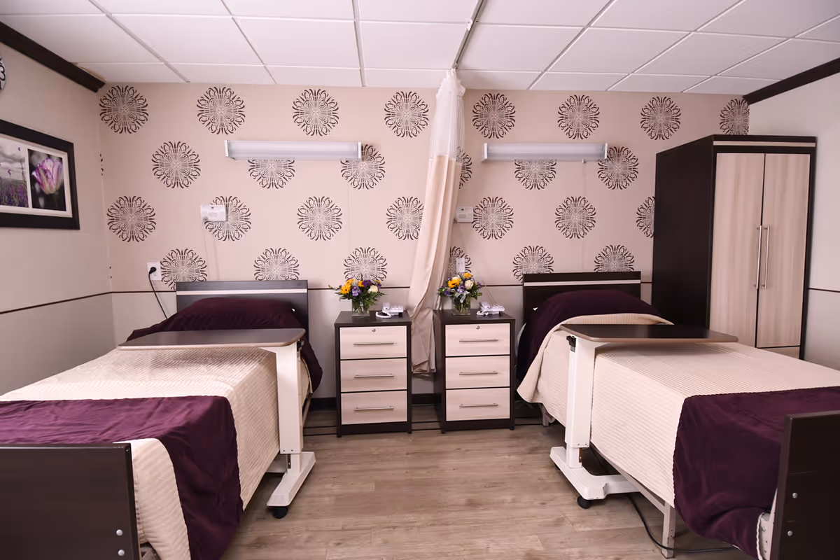 A shared patient room in a rehabilitation and nursing facility with two hospital beds separated by a privacy curtain. Each bed has beige bedding with a dark purple blanket. Between the beds are two bedside tables with drawers, each holding a vase of flowers and a telephone. The walls have a patterned wallpaper with circular designs, and there is a wardrobe on the right side of the room. The floor is wooden, and the ceiling has white tiles.