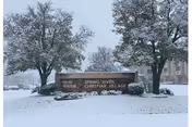 Snow-covered entrance sign for Spring River Christian Village with trees and a building partially visible in the background during a snowy day.