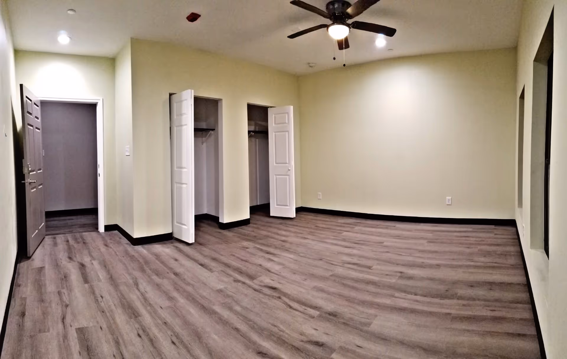 Empty room with light yellow walls, wood flooring, two open closets with white doors, a ceiling fan with light, and an open door leading to another room.