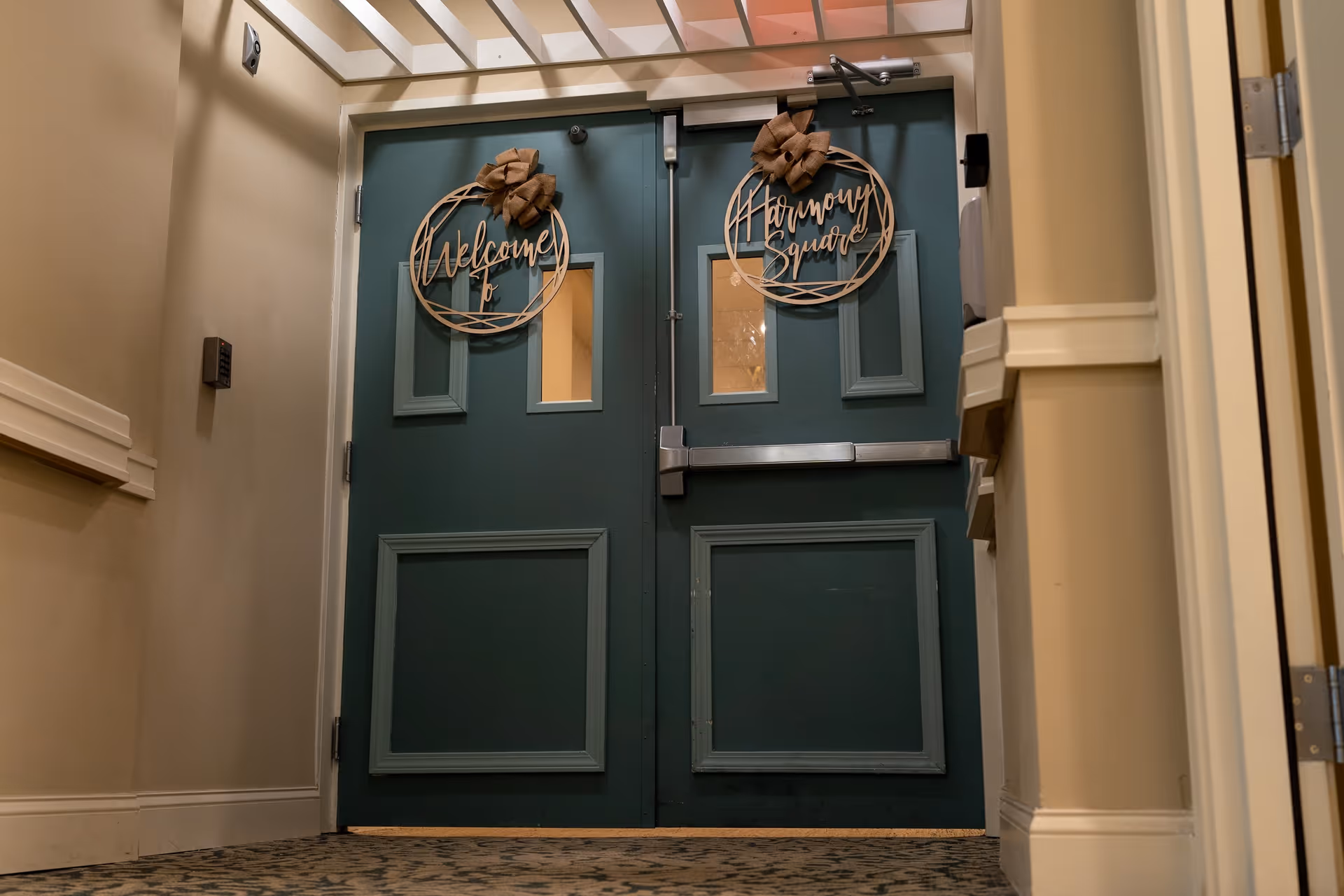Double green doors with small rectangular windows near the top, each decorated with a round wooden wreath. The left wreath says 'Welcome to' and the right wreath says 'Harmony Square'. The doors are set in a beige hallway with patterned carpet and a keypad on the left wall.