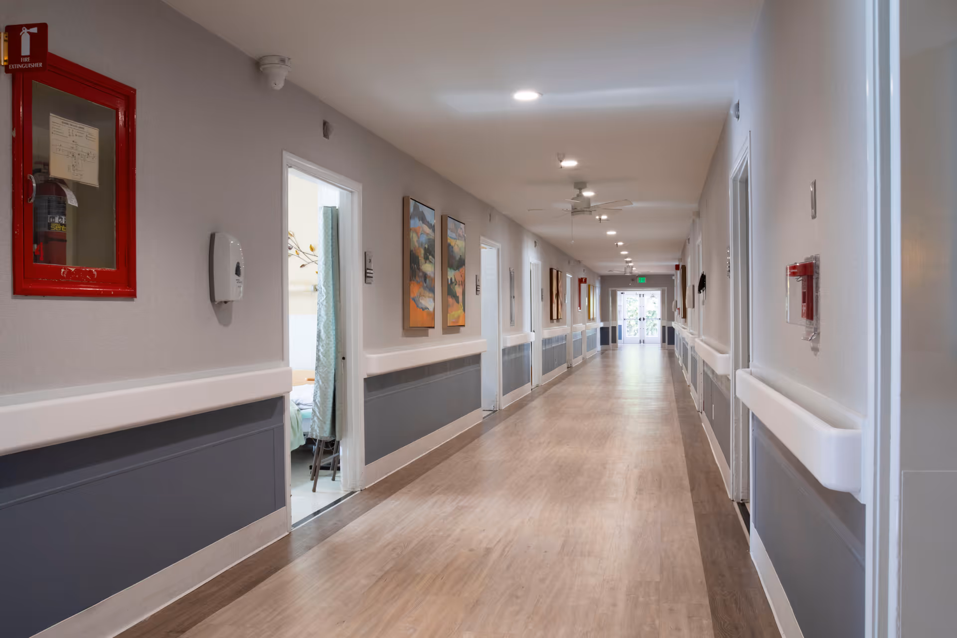 Long, well-lit interior hallway of a senior living facility with handrails, wall art, and open room doors.