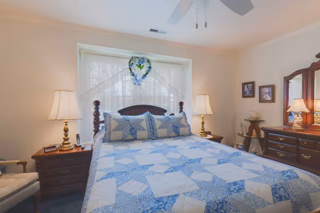 Well-lit bedroom with a large bed covered by a blue star-patterned quilt, bedside tables with lamps, and a dresser with mirror.