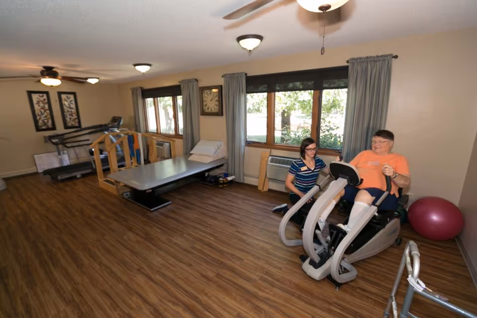 A rehabilitation exercise room with wood flooring, treadmills and therapy equipment, and a staff member assisting an older adult using a recumbent exercise machine near windows.