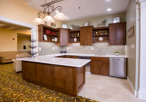 A modern kitchen area with wooden cabinets, a white countertop island, and a stainless steel dishwasher. The backsplash features small mosaic tiles, and there are decorative items and plants on the shelves and cabinet tops. A light fixture with three lamps hangs above the island, and part of a living room with beige sofas is visible in the background.