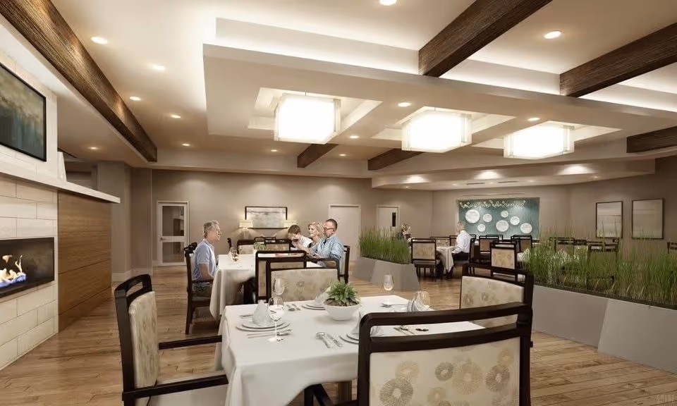 A spacious, well-lit dining room in a senior living facility with several tables covered in white tablecloths, set with glassware and silverware. There are a few people seated and dining, and the room features modern ceiling lights, wooden beams, a fireplace, and decorative plants in planters dividing the space.