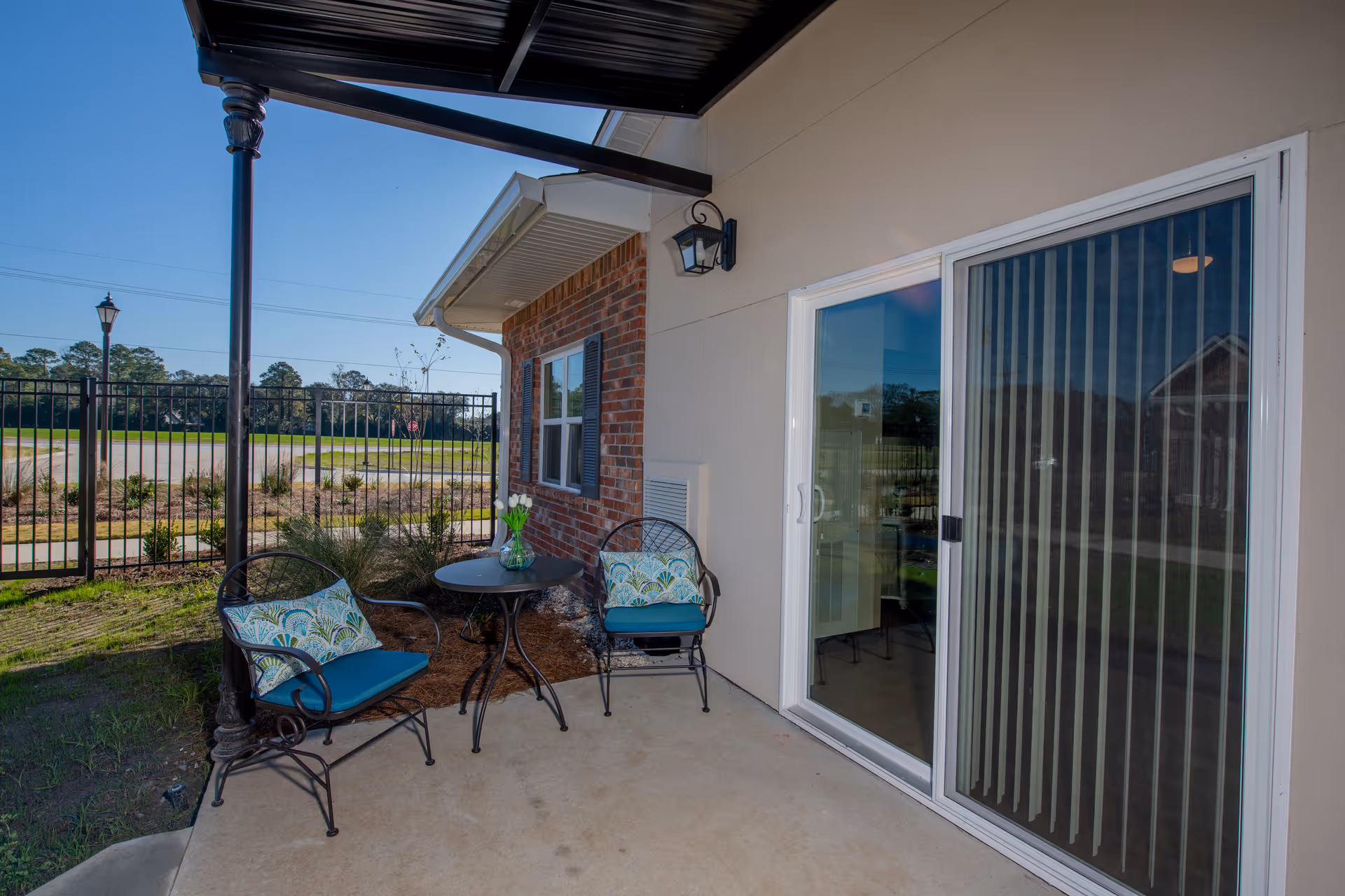Outdoor patio area with two black metal chairs with blue and patterned cushions, a small round black metal table with a vase of white flowers, a sliding glass door with vertical blinds, a brick and beige exterior wall, and a black metal fence with a lamp post in the background under a clear blue sky.