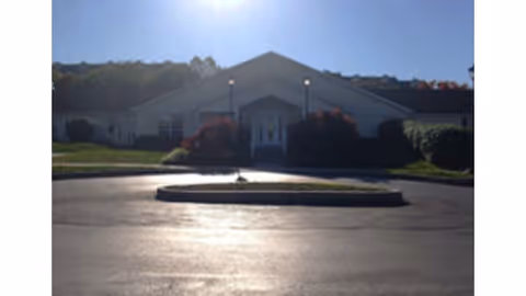 Front exterior view of a single-story building with a peaked roof, surrounded by bushes and greenery, under a clear sky with the sun shining brightly above.