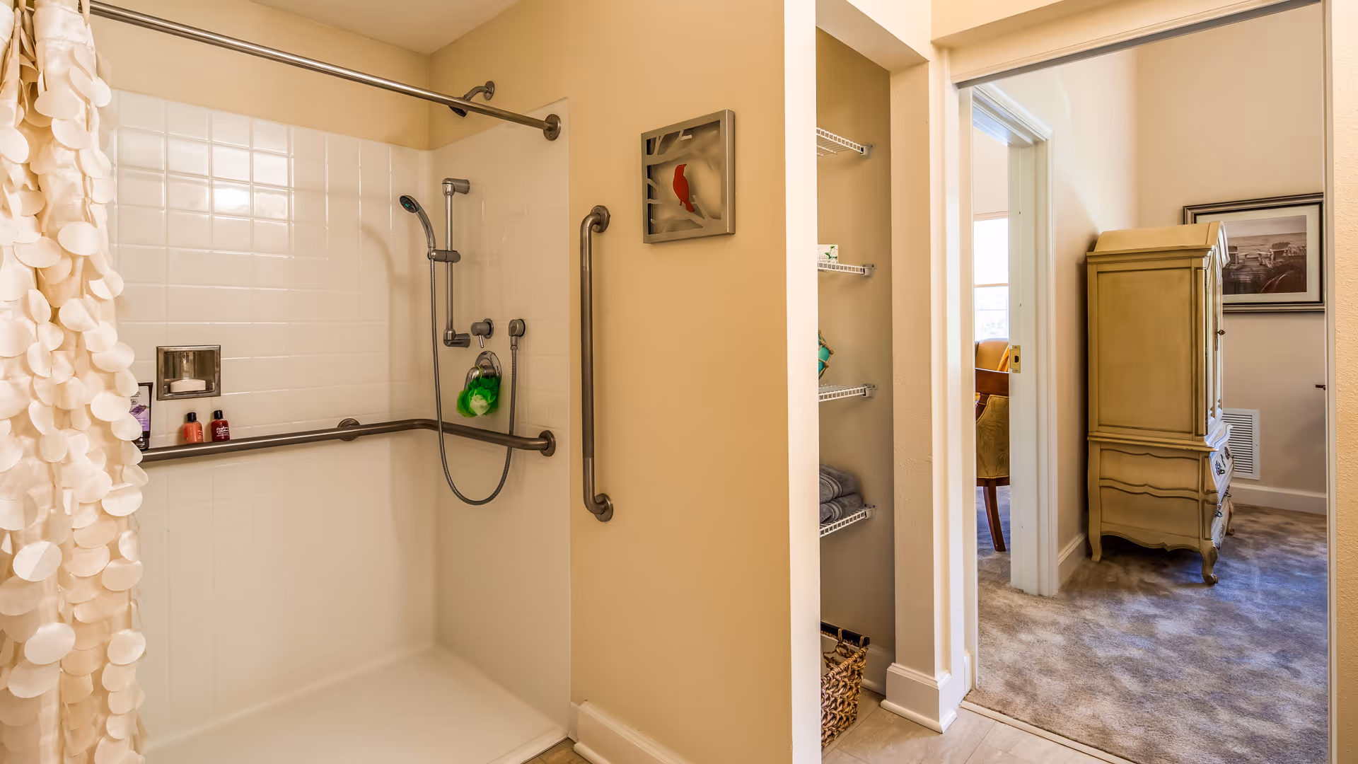 A bathroom with a walk-in shower featuring white tiled walls, a handheld showerhead, and multiple grab bars for accessibility. The shower has a white curtain with circular patterns. Adjacent to the shower is a small open closet with shelves holding towels and other items. Beyond the bathroom, a carpeted room is visible with a wooden cabinet and a framed picture on the wall.