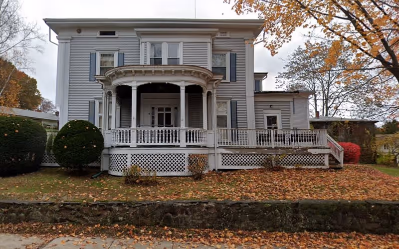 Front exterior view of a two-story white house with a covered porch and decorative railings, surrounded by autumn trees with fallen leaves on the ground.