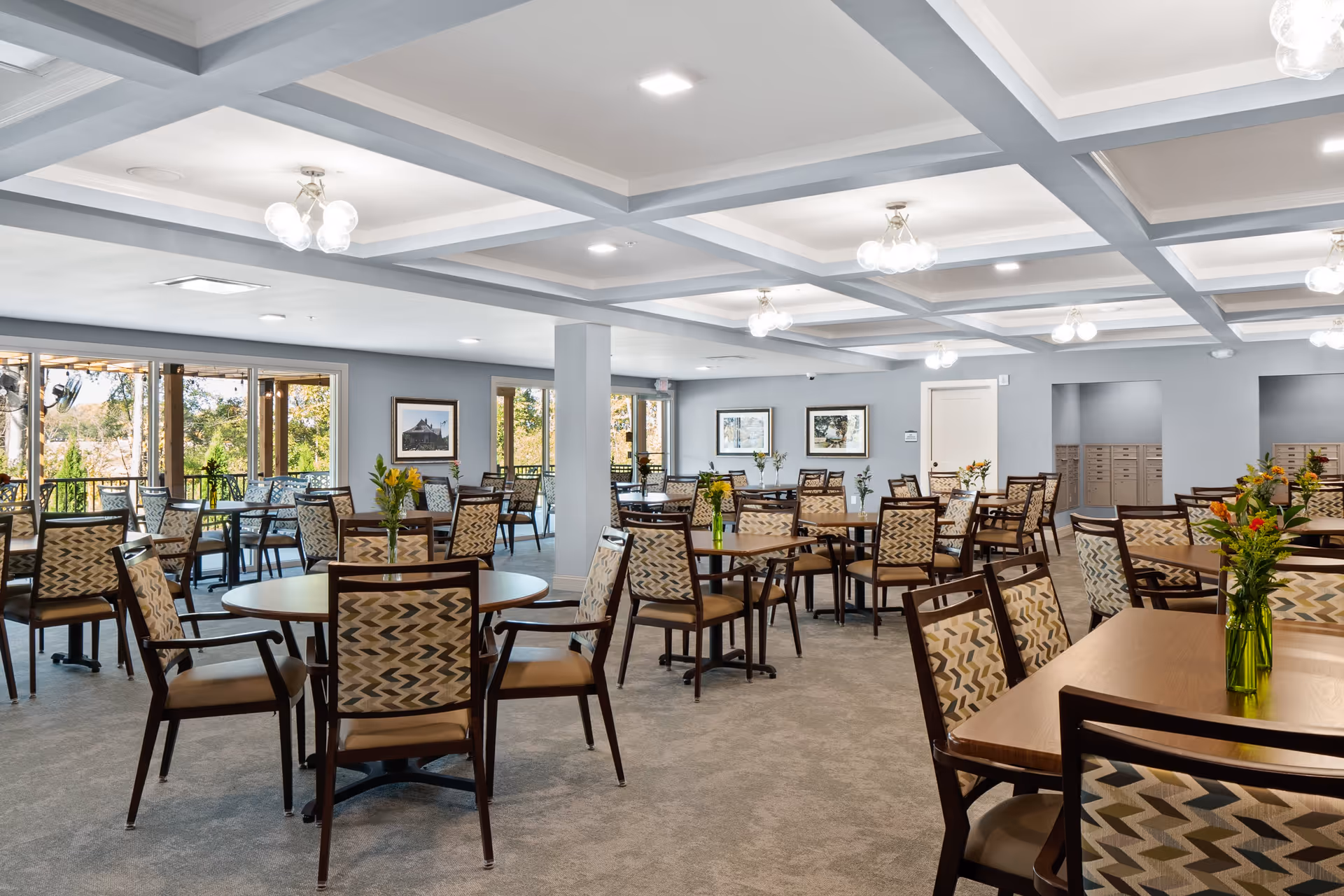 A spacious dining room with multiple round and rectangular wooden tables surrounded by patterned cushioned chairs. Each table has a small vase with fresh flowers. The room has a coffered ceiling with modern light fixtures and large windows letting in natural light, offering a view of greenery outside.