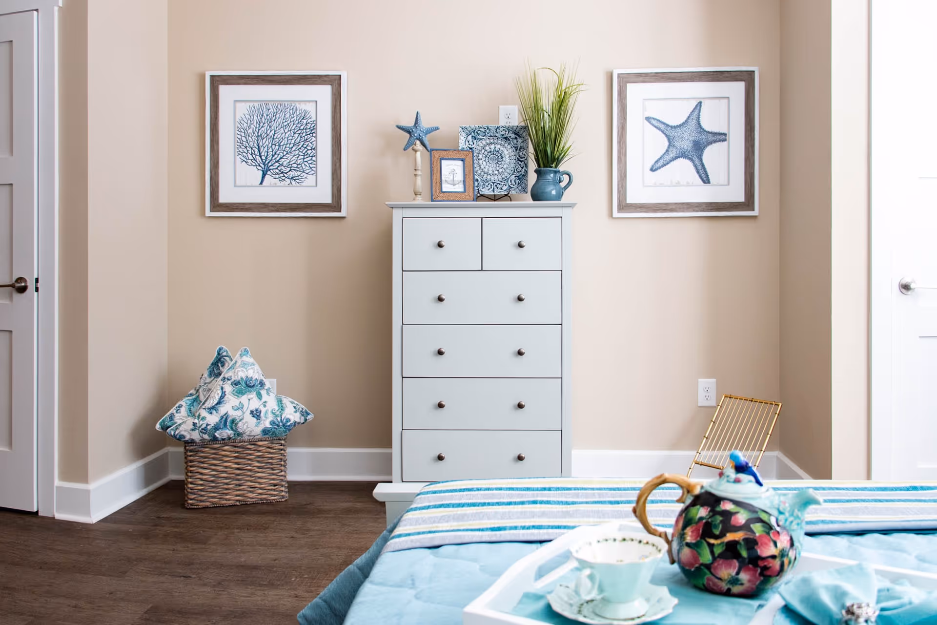 A bedroom corner featuring a white chest of drawers with decorative items on top, including a blue starfish, framed pictures, a decorative plate, and a vase with green plants. On the wall above the chest are two framed pictures of sea life, one of coral and one of a starfish. To the left, a wicker basket holds two blue and white patterned pillows. In the foreground, part of a bed with a blue quilt and a tray holding a floral teapot and a teacup is visible.