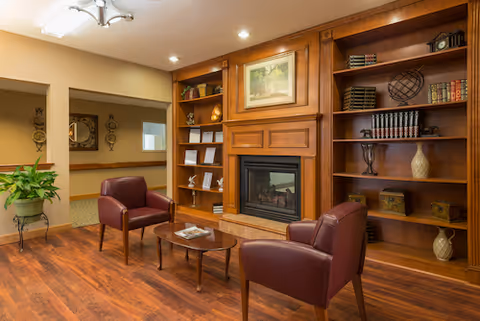 A cozy living room area with two maroon leather armchairs facing a wooden coffee table. Behind the chairs is a wooden built-in bookshelf with various decorative items and books, and a fireplace in the center with a framed painting above it. The room has wooden flooring and warm lighting.