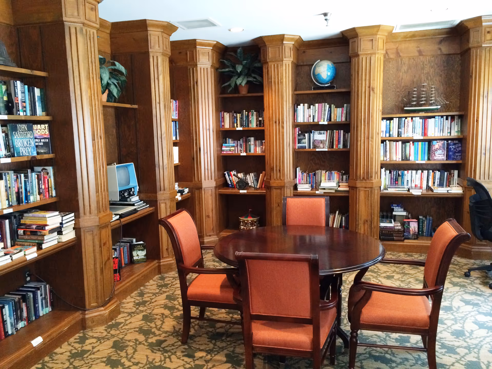 A cozy library room with wooden bookshelves filled with books and decorative items, including a globe and a model ship. In the center is a round wooden table surrounded by four orange upholstered chairs. The room has a patterned carpet and wooden paneling on the walls.