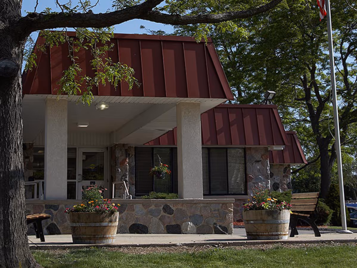 Exterior view of a building with a red sloped roof, stone and concrete walls, and a covered entrance supported by columns. There are two large wooden planters with colorful flowers on either side of the entrance, a bench to the right, and trees surrounding the area under a clear blue sky.