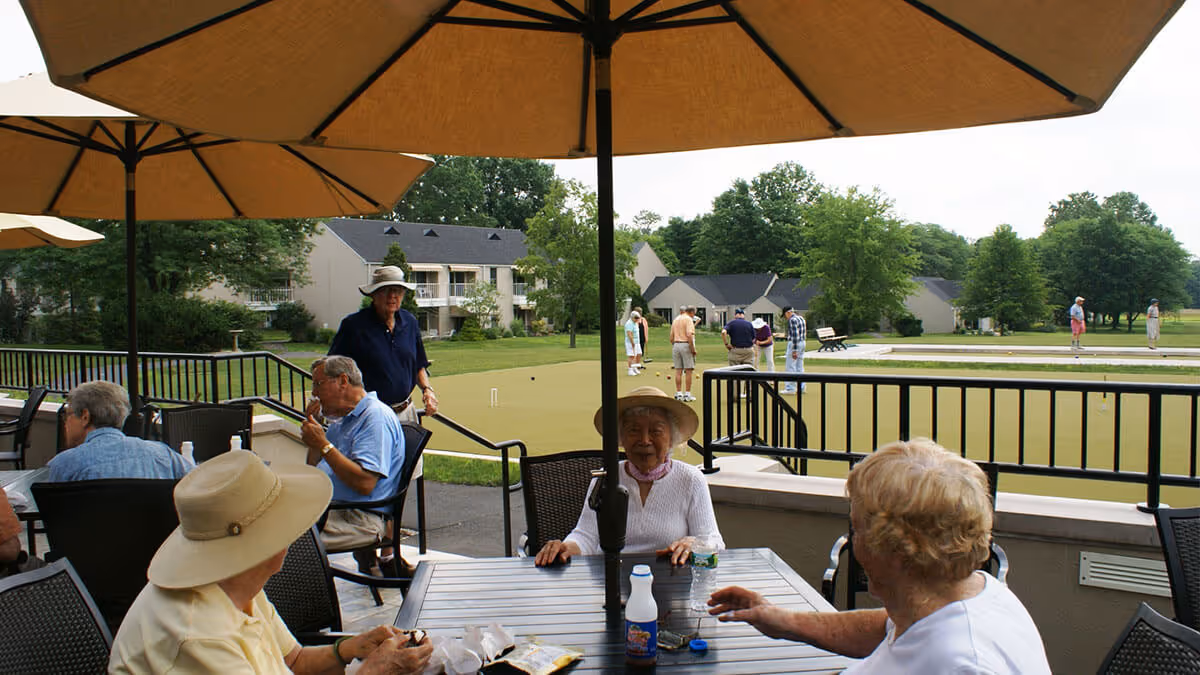 Older adults sit at patio tables under large umbrellas beside a lawn bowling green with residential buildings in the background.
