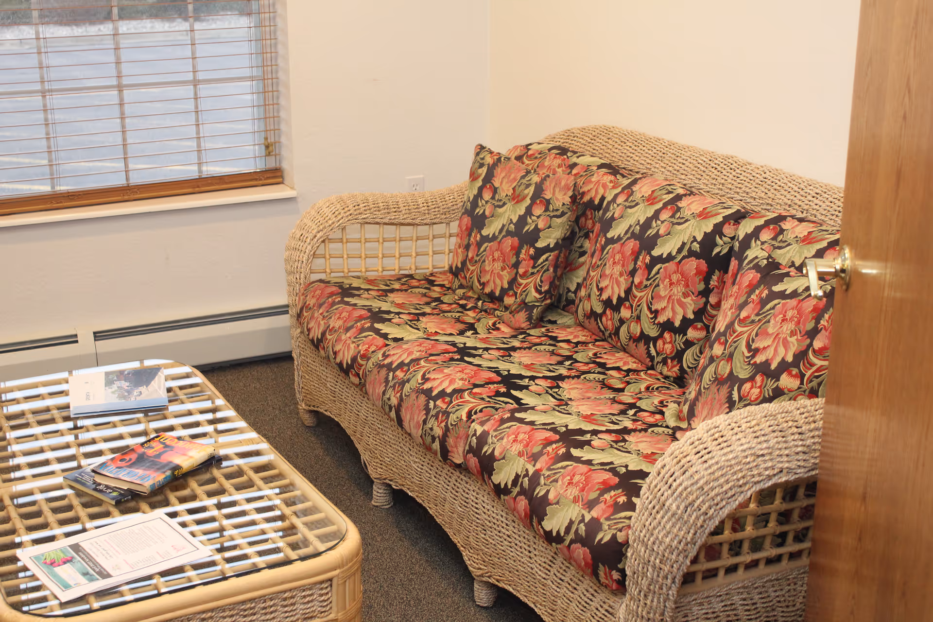 A cozy living room area featuring a wicker sofa with floral patterned cushions in shades of red, green, and black. In front of the sofa is a matching wicker coffee table with a glass top, holding several magazines and brochures. A window with wooden blinds is visible on the left side, and a wooden door is partially open on the right.