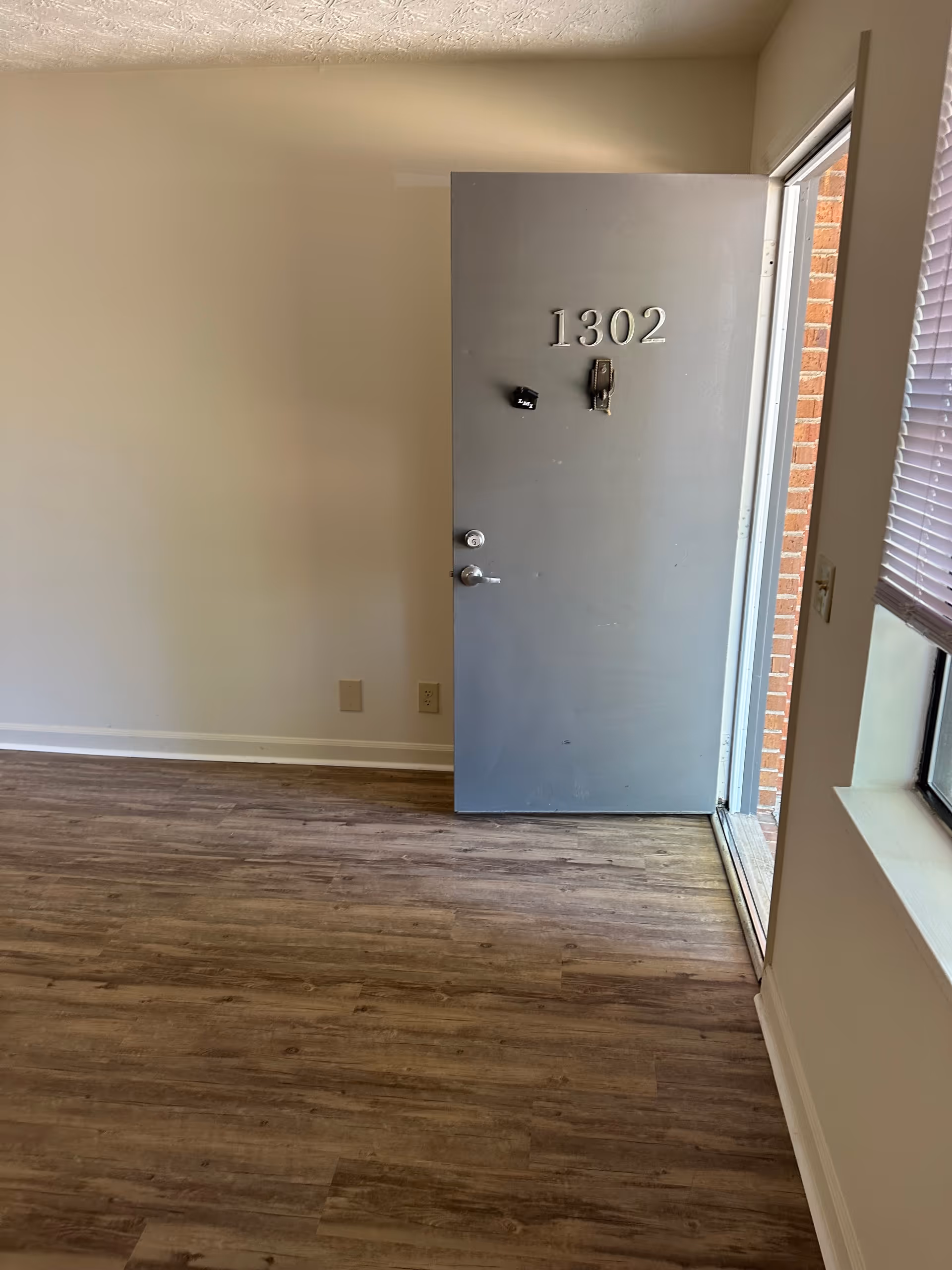 An open gray door with the number 1302 on it, leading to the outside. The floor is wood-patterned, and there is a window with blinds partially visible on the right side. The walls are plain and light-colored.