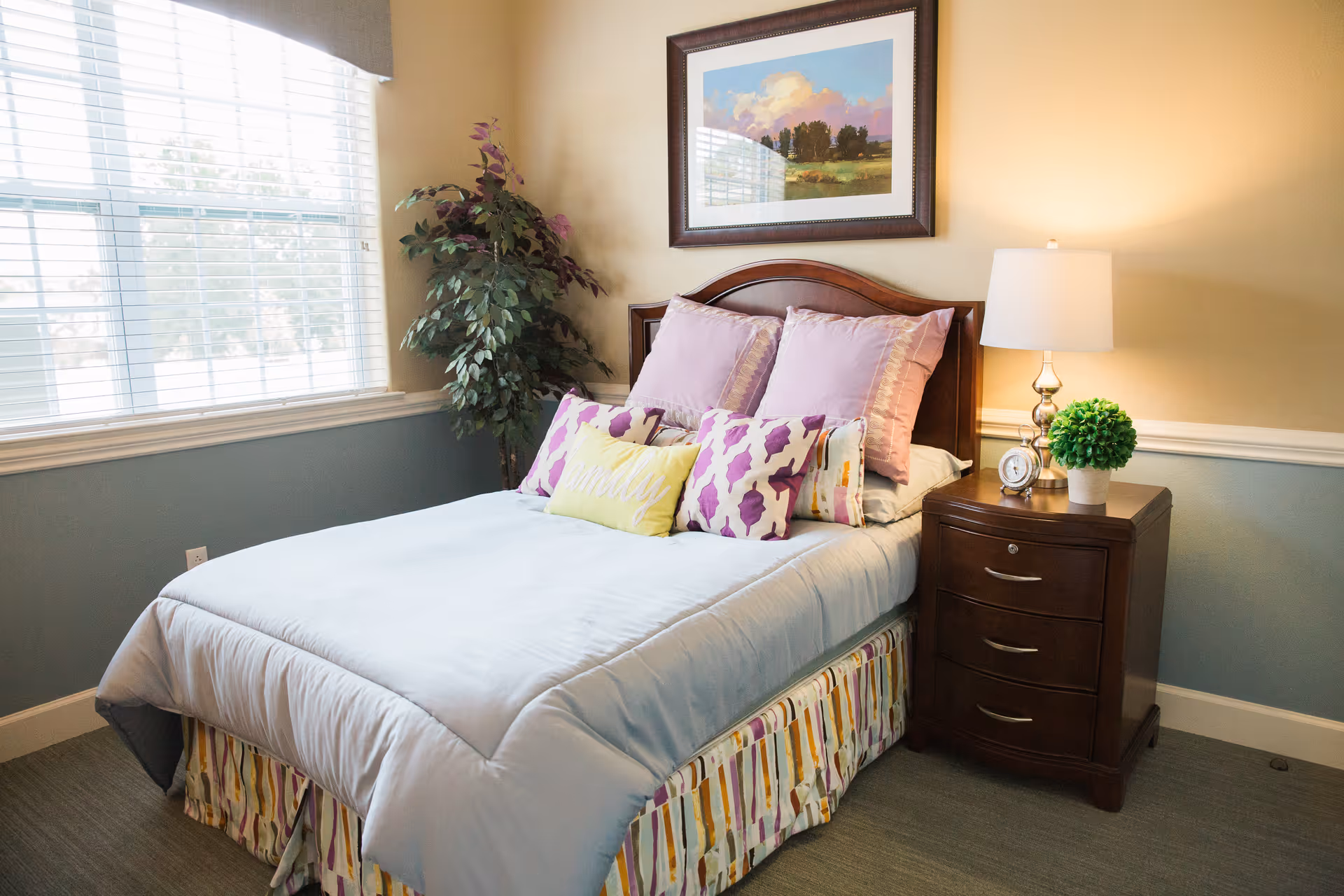 Sunlit bedroom with a neatly made twin bed, patterned pillows, a wooden nightstand with lamp and plant, and framed artwork above the headboard.