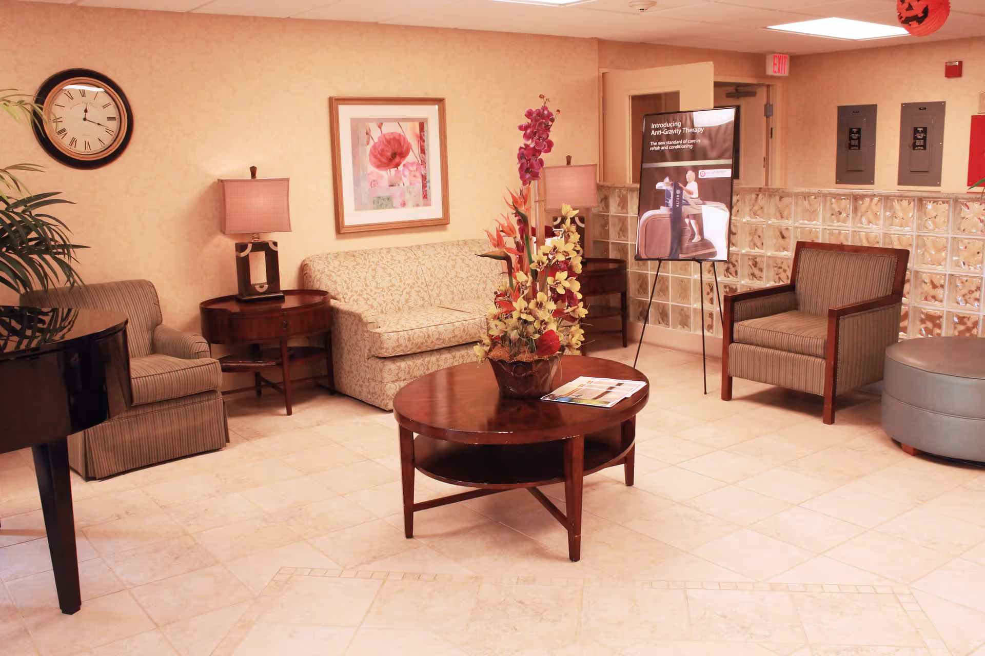 A cozy seating area in a senior living facility with a beige patterned sofa, two striped armchairs, a round wooden coffee table with a floral arrangement, a side table with a lamp, a wall clock, a framed floral painting, and a promotional stand about Anti-Gravity Therapy. The floor is tiled and the walls are light-colored.