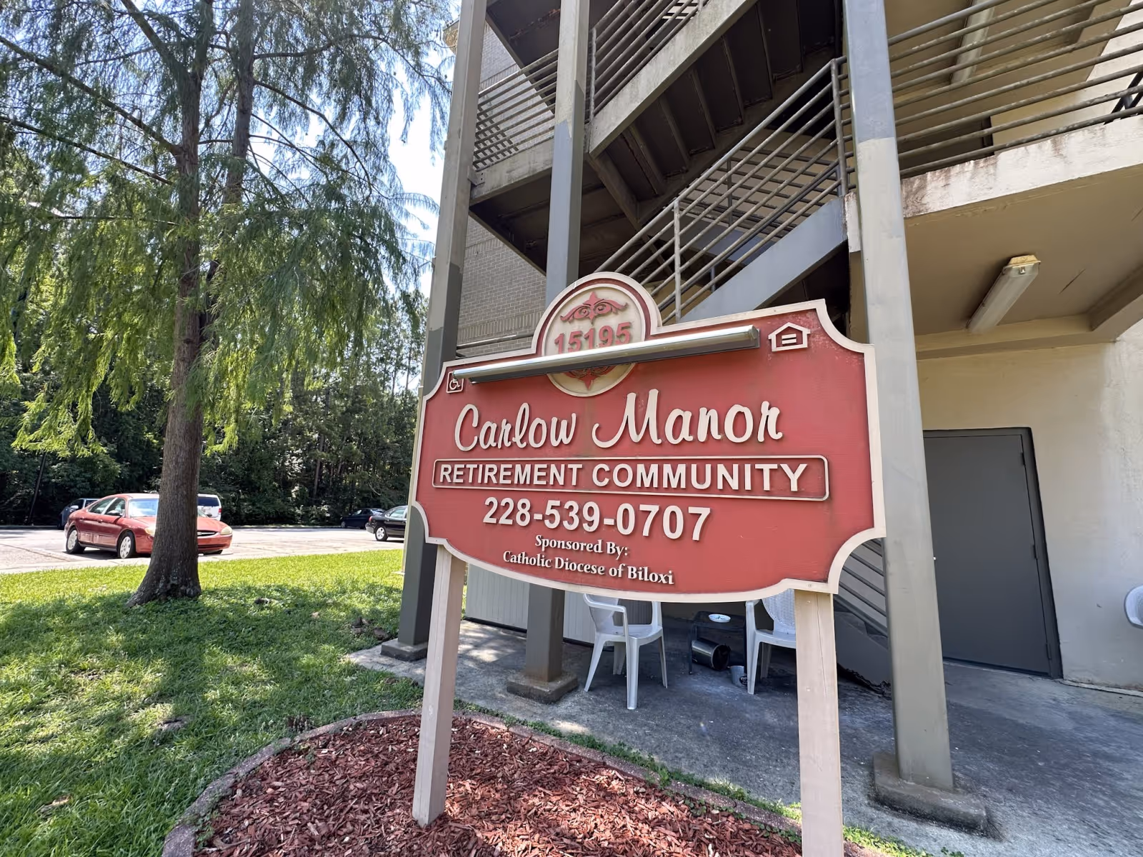 Red Carlow Manor Retirement Community sign in front of a multi-story building with exterior stairs and a few white chairs nearby.