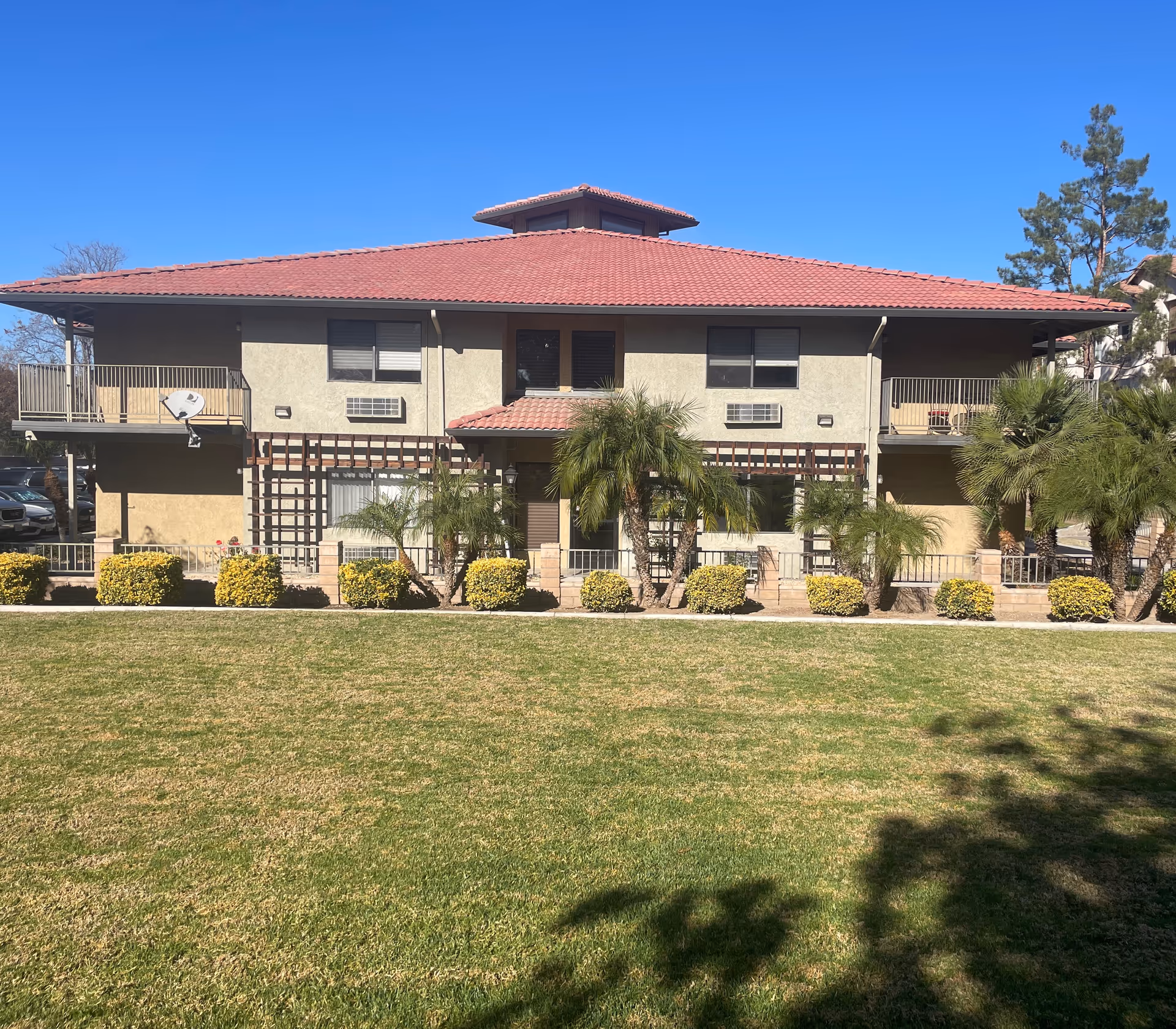 Exterior view of a two-story building with a red tiled roof, beige walls, balconies on both sides, and several windows. The building is surrounded by neatly trimmed bushes, palm trees, and a well-maintained green lawn under a clear blue sky.