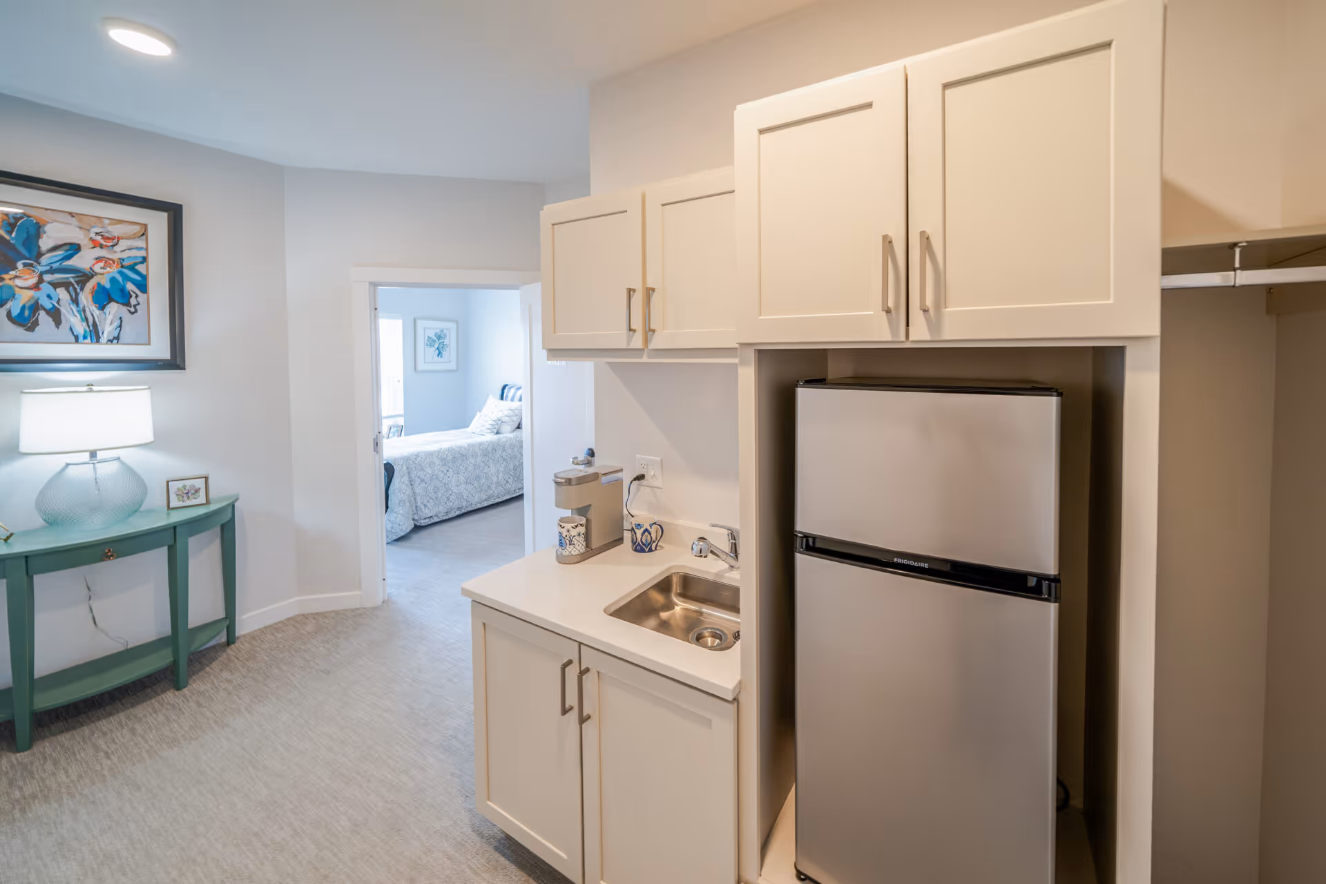 Interior view of a senior living facility unit showing a small kitchenette with white cabinets, a stainless steel mini refrigerator, and a sink. To the left, there is a green console table with a lamp and a framed artwork on the wall. In the background, a bedroom with a bed and framed pictures is visible through an open doorway.