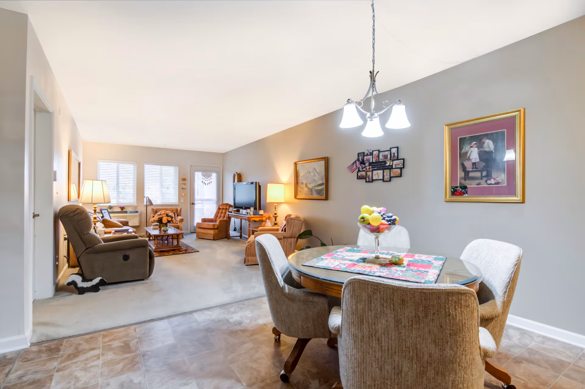 A cozy living and dining area in a retirement community. The foreground shows a round dining table with four cushioned chairs and a centerpiece bowl of fruit. The background features a living room with multiple armchairs, a coffee table, a TV on a stand, and framed pictures on the walls. The room is well-lit with natural light coming through windows and a glass door.