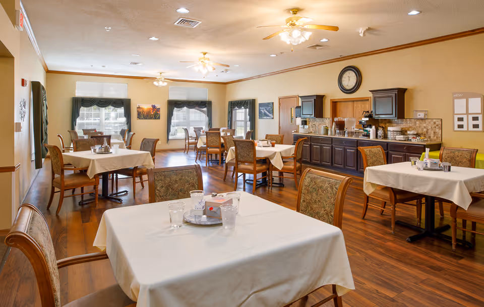 Well-lit dining room with multiple tables covered in white tablecloths, chairs, and a serving counter along the back wall.