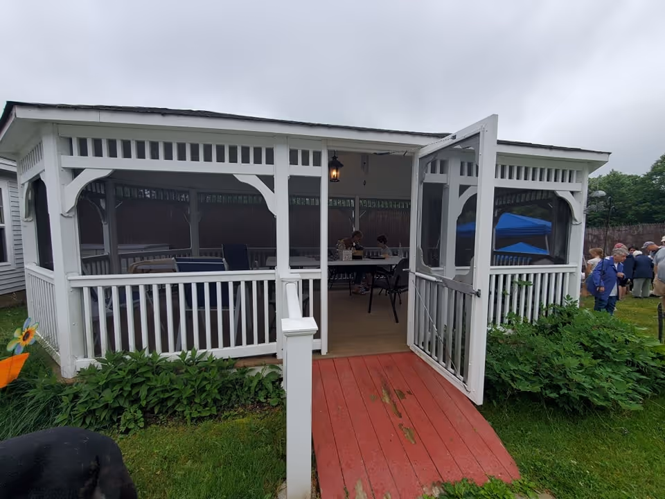 A white screened-in gazebo with an open door and a red wooden ramp leading up to it. Inside, there are tables and chairs with people seated. The gazebo is surrounded by green grass and plants, and a group of people is gathered outside on the right side under a cloudy sky.