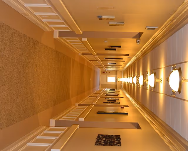 A long, well-lit hallway with beige walls and carpeted floor. The ceiling has a row of evenly spaced wall-mounted light fixtures on one side. The hallway features white wainscoting and decorative wall art along the walls, leading to a door with a window at the far end.