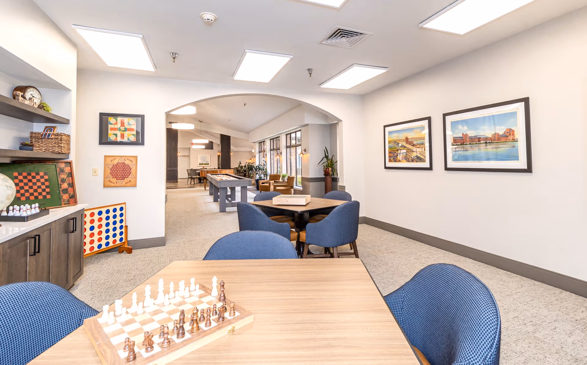 A bright and spacious senior living common area with tables and blue chairs arranged for games. A chessboard is set up on the nearest table. The room features a shuffleboard table in the background, large windows letting in natural light, framed artwork on the walls, and shelves with board games and decorative items.
