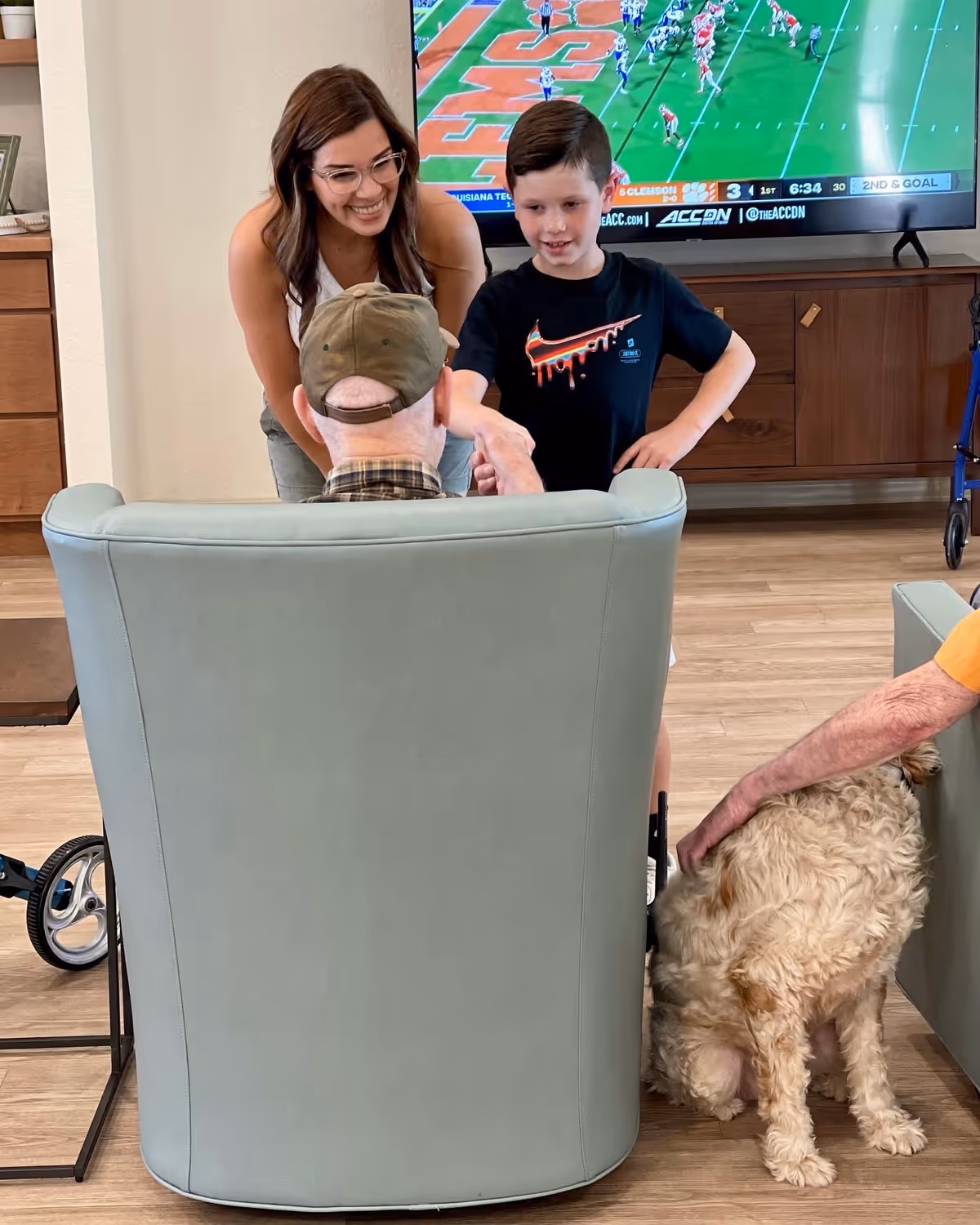 A young boy and a woman smiling and interacting with an elderly man seated in a light green armchair in a living room. A dog is being petted by another person sitting nearby. A large flat-screen TV showing a football game is mounted on the wall in the background.