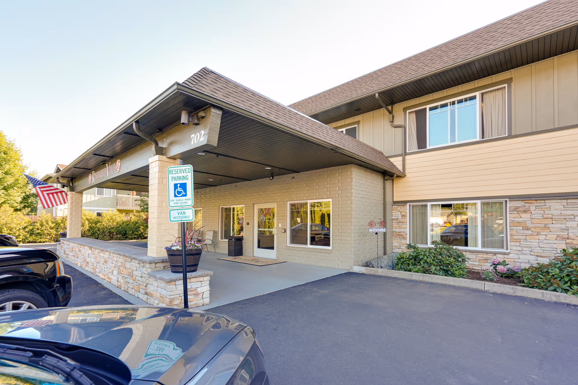 Covered main entrance of a two-story senior living building with an accessible parking sign, planter, and an American flag.