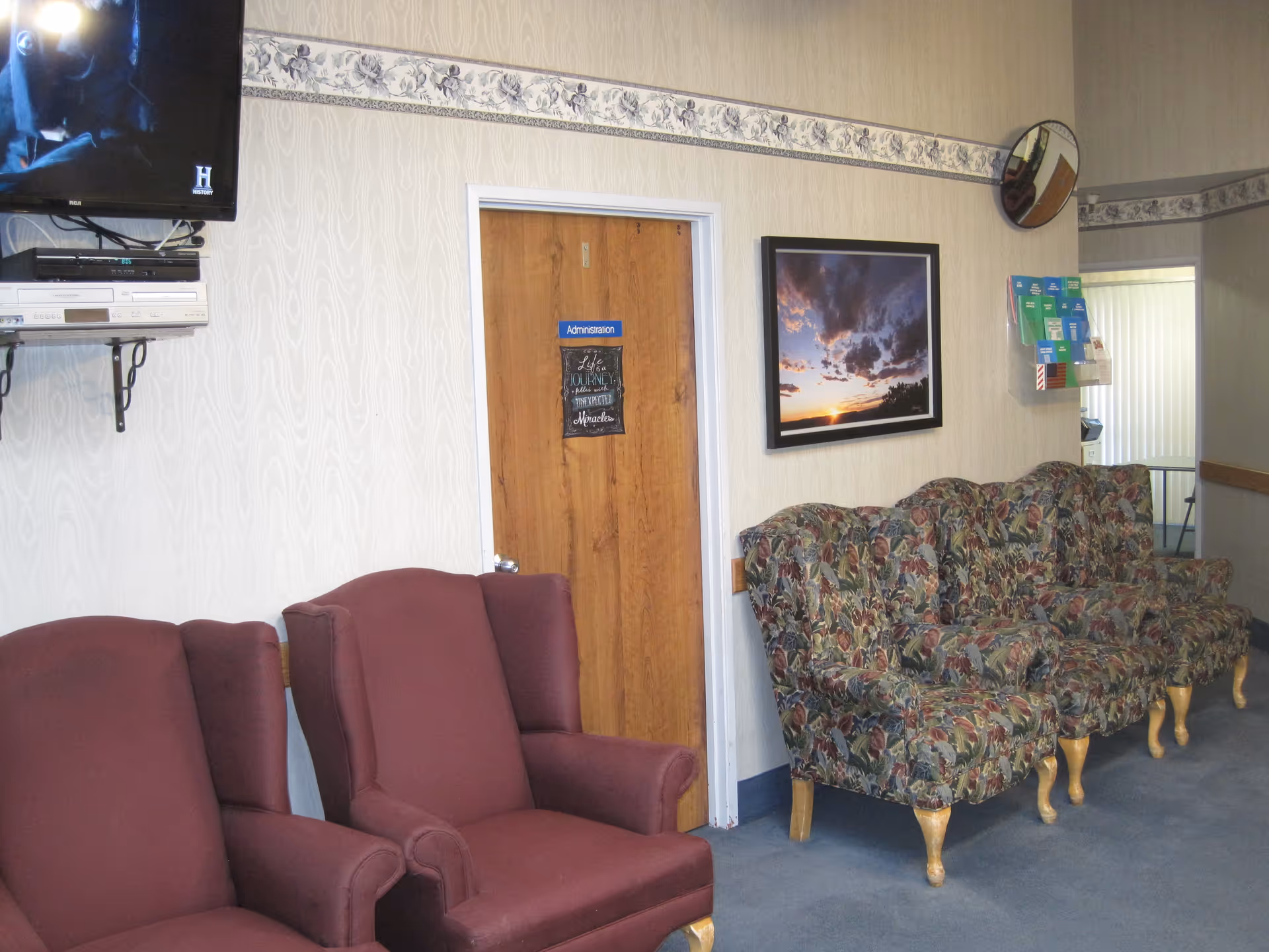 Waiting room with maroon armchairs and floral-upholstered chairs facing a wood door labeled 'Administration', a framed sunset picture, and a wall-mounted TV.