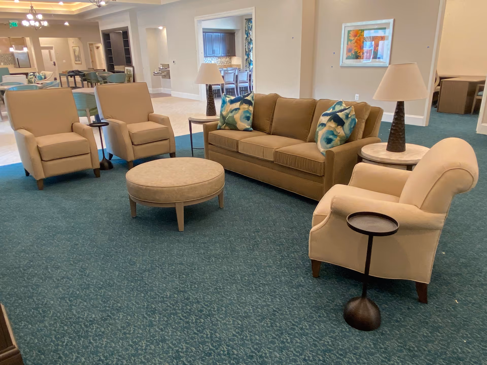 Communal lounge with a sofa, armchairs, round ottoman and side tables on teal carpet in a senior living facility.