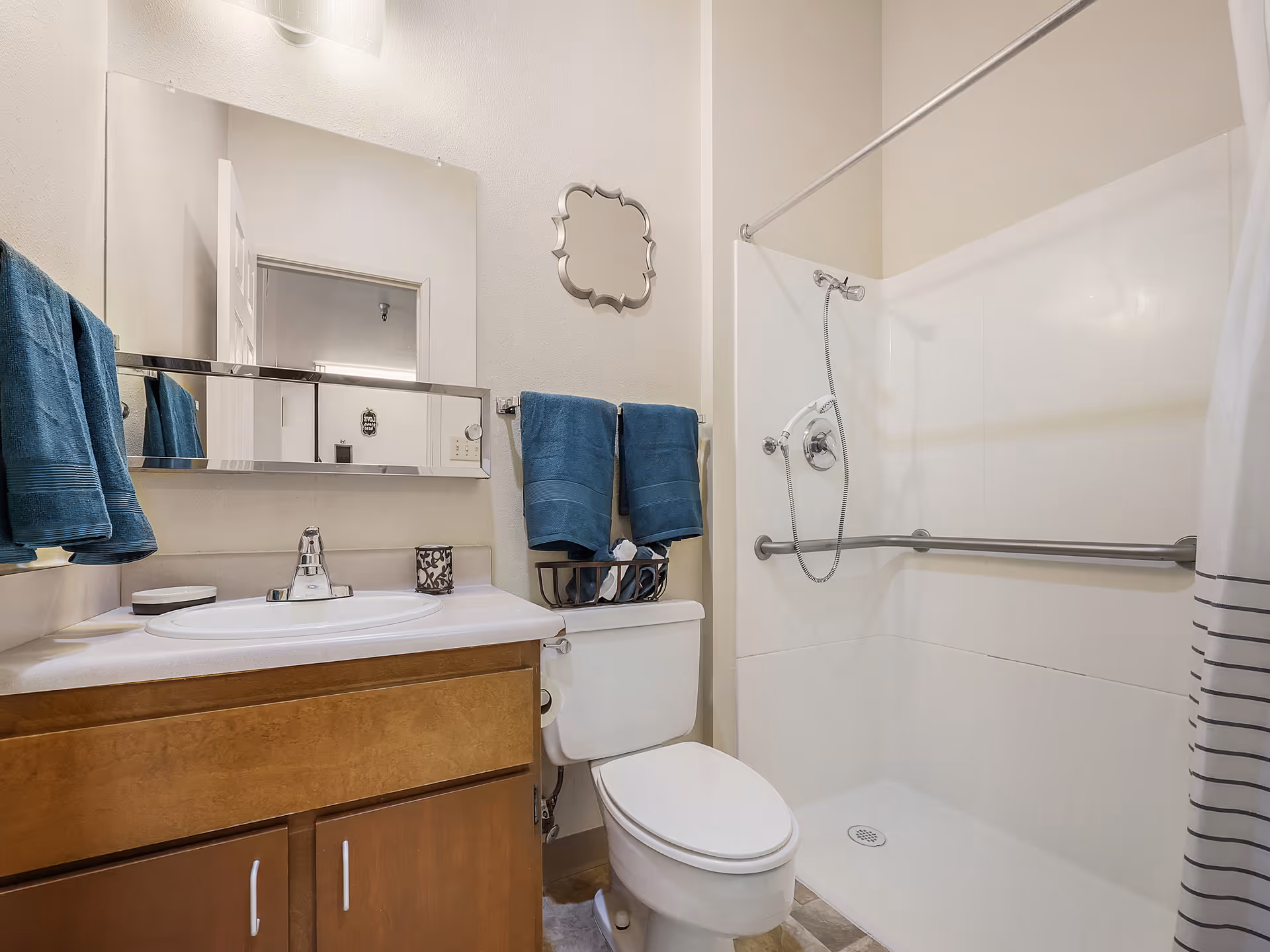 A clean bathroom with a wooden vanity cabinet, white sink, and a large mirror above it. There are blue towels hanging on towel racks next to a white toilet. The shower area has a handheld showerhead, grab bars, and a white shower curtain with gray stripes.