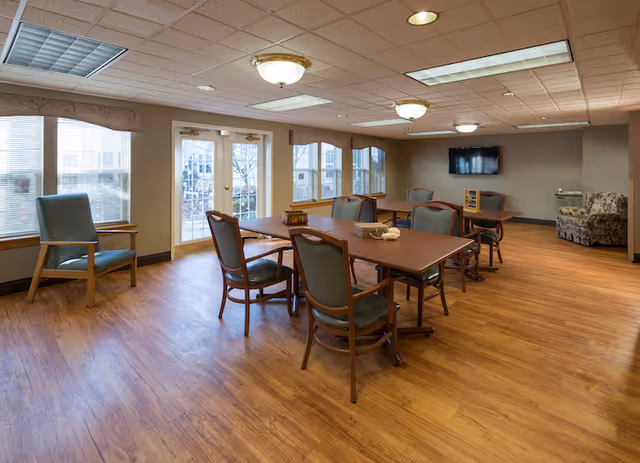 A well-lit room with wooden flooring featuring several tables and chairs arranged for group seating. Large windows and glass doors allow natural light to enter. A flat-screen TV is mounted on the far wall, and there is a patterned armchair in the corner.