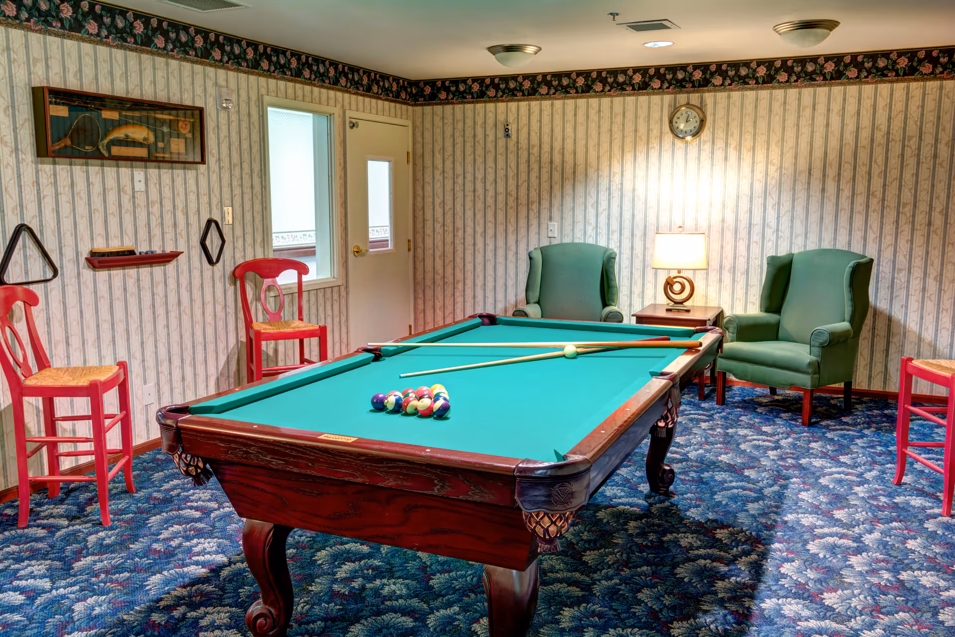 A pool table sits in a cozy carpeted game room with green armchairs, red wooden chairs, floral wallpaper and a side table lamp.